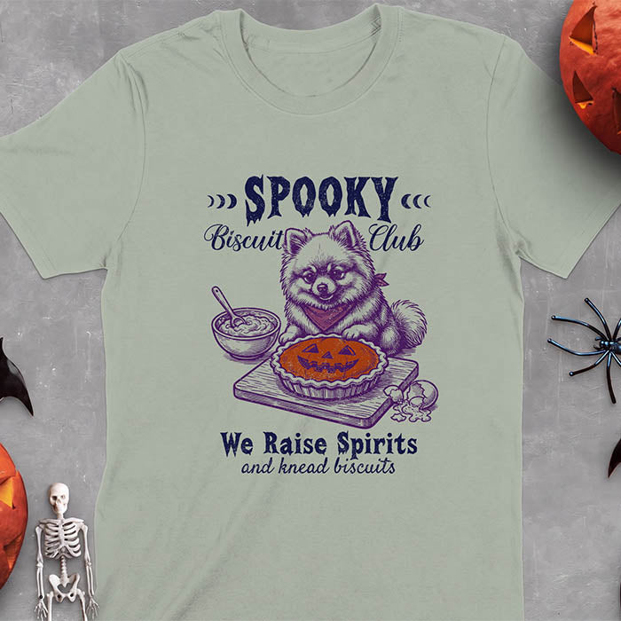 Sage t-shirt with a Halloween graphic of a Pomeranian wearing a plaid bandana sitting behind a jack-o'-lantern pumpkin pie on a cutting board with a mixing bowl, spoon and a cracked egg nearby and the text Spooky Biscuit Club, We Raise Spirits and knead biscuits