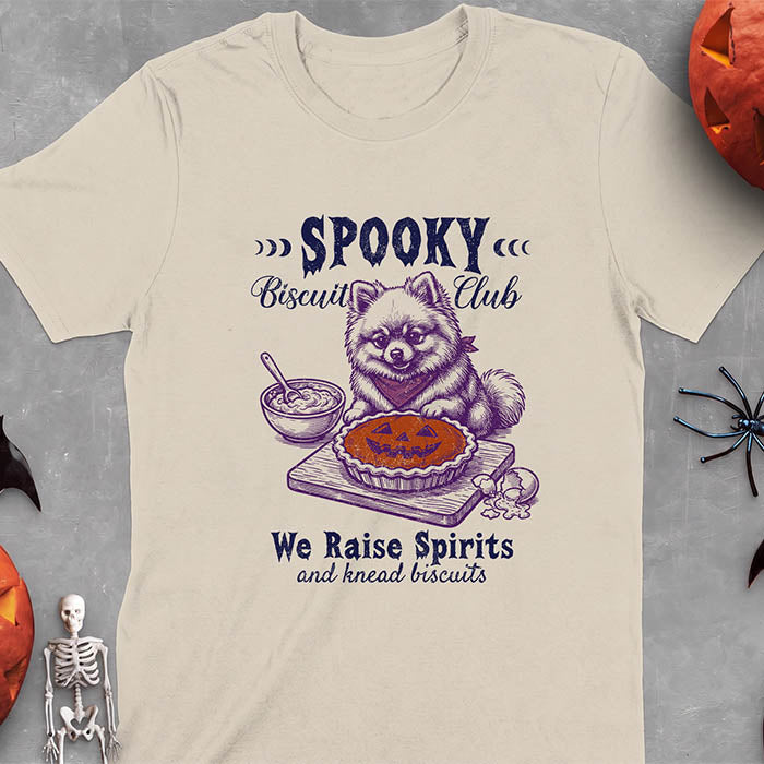 Sand t-shirt with a Halloween graphic of a Pomeranian wearing a plaid bandana sitting behind a jack-o'-lantern pumpkin pie on a cutting board with a mixing bowl, spoon and a cracked egg nearby and the text Spooky Biscuit Club, We Raise Spirits and knead biscuits