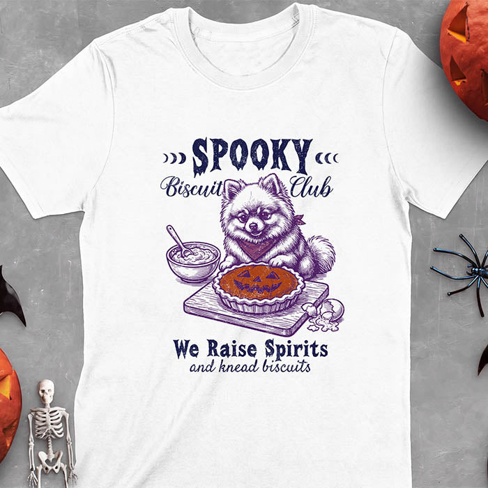 White t-shirt with a Halloween graphic of a Pomeranian wearing a plaid bandana sitting behind a jack-o'-lantern pumpkin pie on a cutting board with a mixing bowl, spoon and a cracked egg nearby and the text Spooky Biscuit Club, We Raise Spirits and knead biscuits