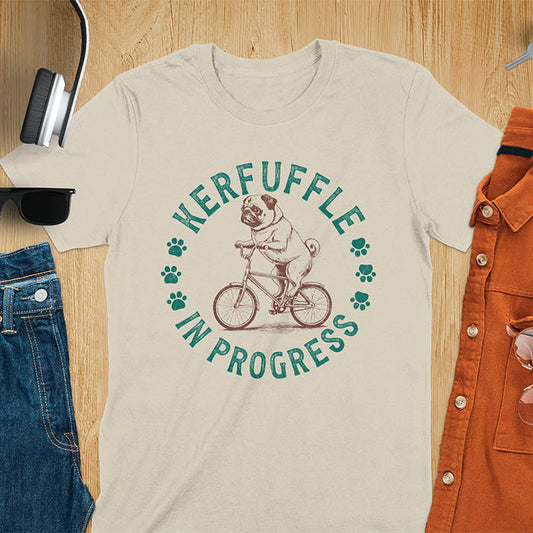 Pug on bicycle sketch-style graphic with Kerfuffle In Progress text and paw prints on sand color t-shirt