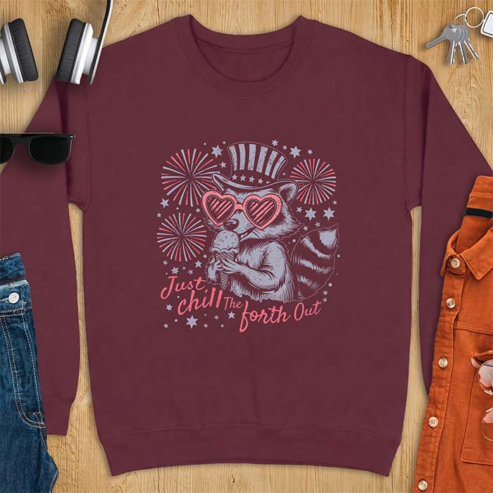 Maroon sweatshirt with a graphic of a raccoon in a striped top hat and heart-shaped sunglasses eating an ice cream cone in front of fireworks and stars, printed with the text Just Chill The Forth Out