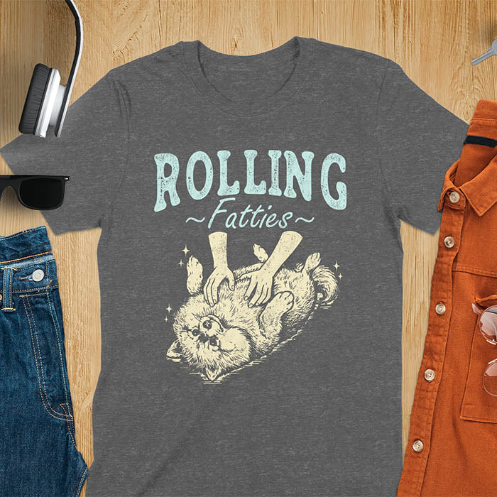 Dark heather t-shirt with vintage-inspired illustration. Retro graphic features fluffy Pomeranian rolling on back with 'Rolling Fatties' text in distressed typography. Quirky chubby dog meme design.