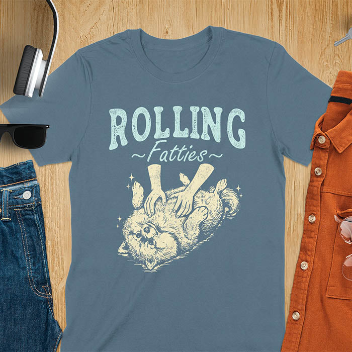Indigo blue t-shirt with vintage-inspired illustration. Retro graphic features fluffy Pomeranian rolling on back with 'Rolling Fatties' text in distressed typography. Quirky chubby dog meme design.
