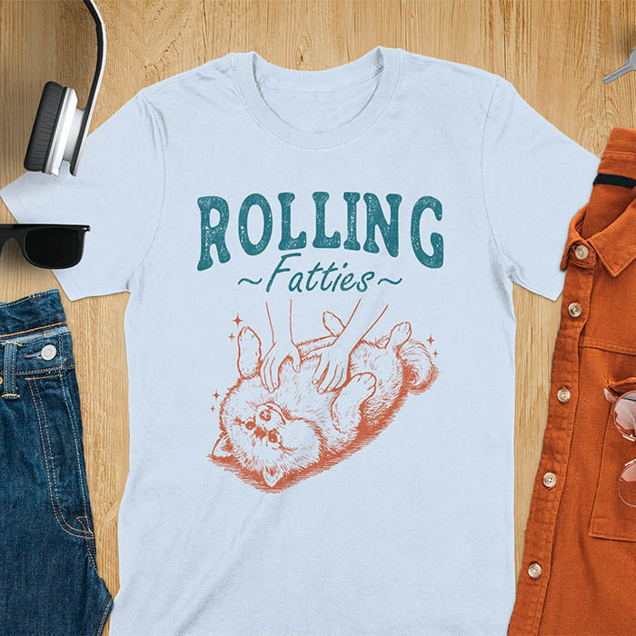 Light blue t-shirt with vintage-inspired illustration. Retro graphic features fluffy Pomeranian rolling on back with 'Rolling Fatties' text in distressed typography. Quirky chubby dog meme design.