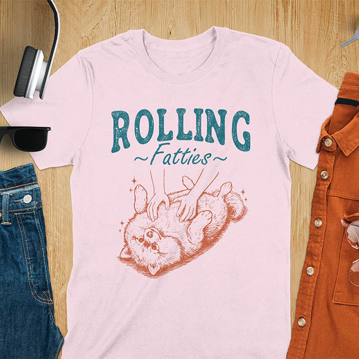 Light pink t-shirt with vintage-inspired illustration. Retro graphic features fluffy Pomeranian rolling on back with 'Rolling Fatties' text in distressed typography. Quirky chubby dog meme design.