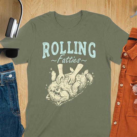 Military Green t-shirt with vintage-inspired illustration. Retro graphic features fluffy Pomeranian rolling on back with 'Rolling Fatties' text in distressed typography. Quirky chubby dog meme design.