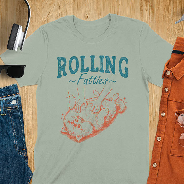 Sage Green t-shirt with vintage-inspired illustration. Retro graphic features fluffy Pomeranian rolling on back with 'Rolling Fatties' text in distressed typography. Quirky chubby dog meme design.