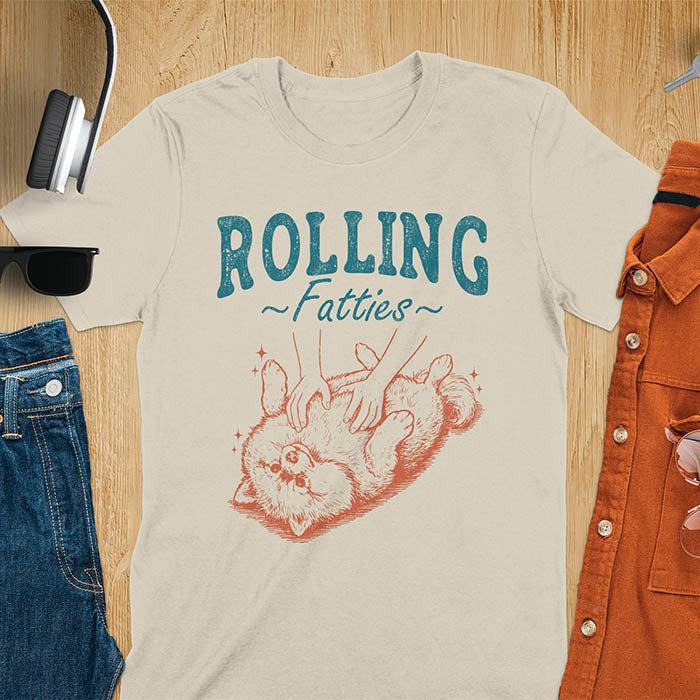 Sand color t-shirt with vintage-inspired illustration. Retro graphic features fluffy Pomeranian rolling on back with 'Rolling Fatties' text in distressed typography. Quirky chubby dog meme design.