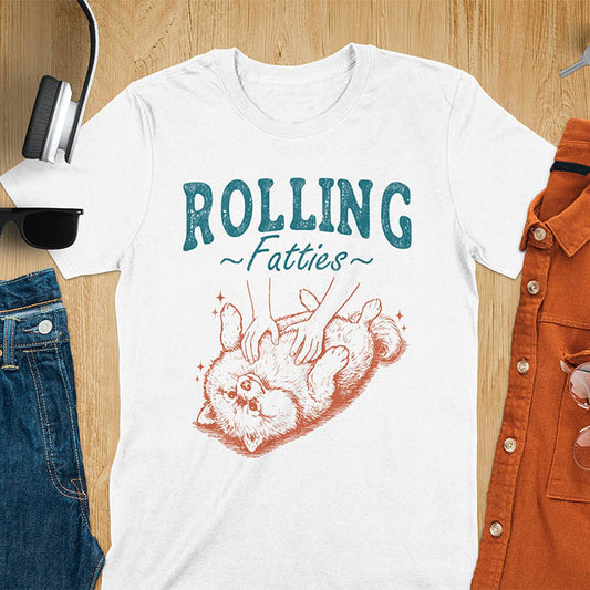 White t-shirt with vintage-inspired illustration. Retro graphic features fluffy Pomeranian rolling on back with 'Rolling Fatties' text in distressed typography. Quirky chubby dog meme design.