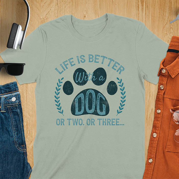 Sage green t-shirt featuring large paw print with text inside surrounded by laurel wreath, text reads Life Is Better With A Dog Or Two Or Three