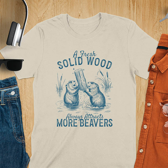 Sand color t-shirt with vintage-style illustration of two beavers in water fighting over a wooden log, featuring text 'A Fresh Solid Wood Always Attracts More Beavers' in rustic lettering 