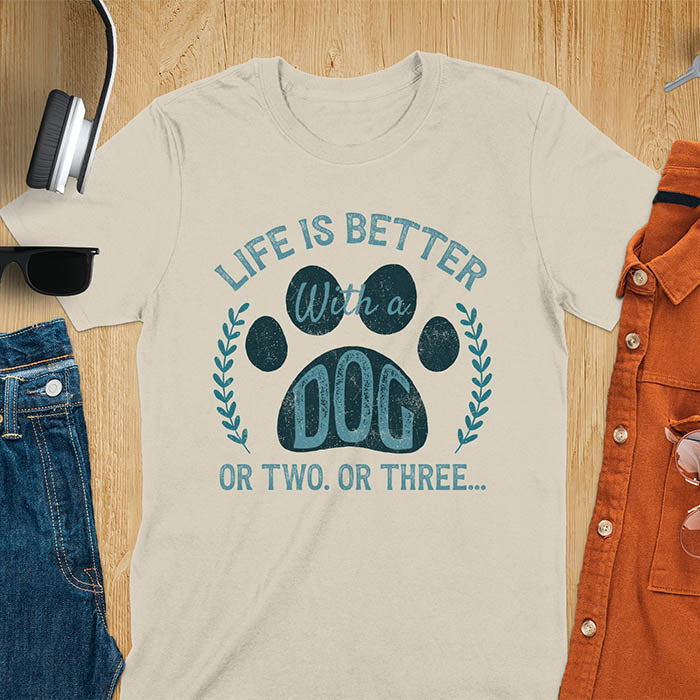 Sand color t-shirt featuring large paw print with text inside surrounded by laurel wreath, text reads Life Is Better With A Dog Or Two Or Three