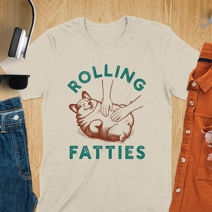 Chubby corgi illustration enjoying tummy rubs with Rolling Fatties text in textured lettering on sand color tee