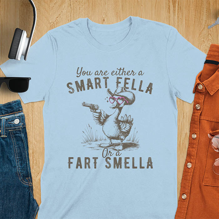 Light blue color tee with A goose wearing heart shaped glasses and holding a pistol. Text reads You are either a Smart Fella or a Fart Smella.