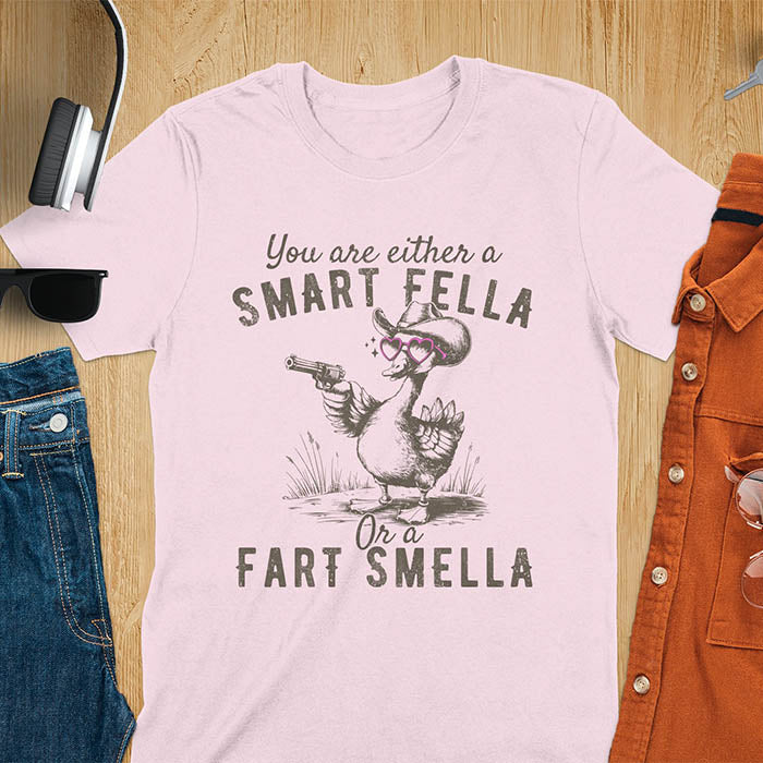 Pink color tee withA goose wearing heart shaped glasses and holding a pistol. Text reads You are either a Smart Fella or a Fart Smella.