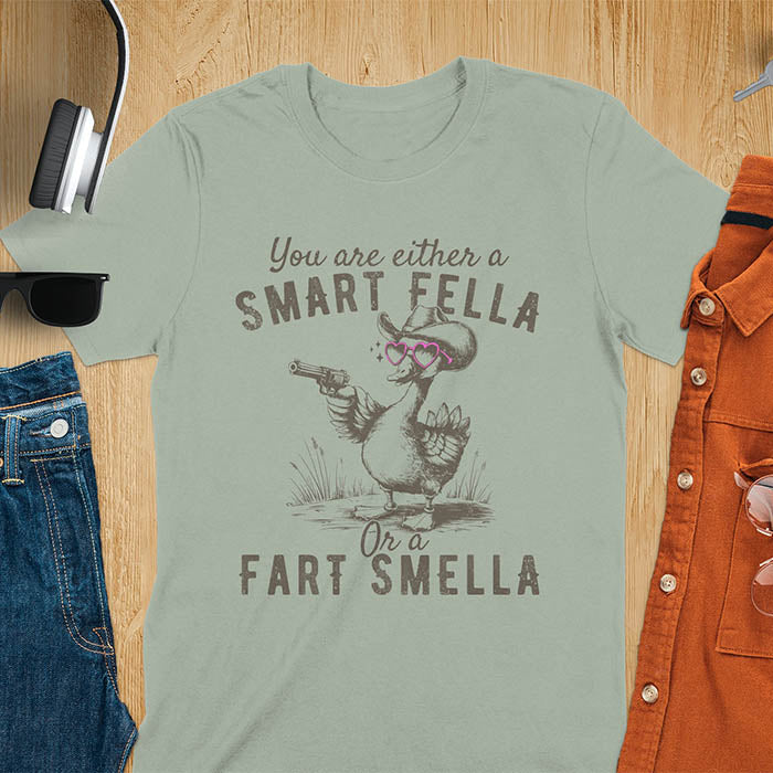 Sage color tee with A goose wearing heart shaped glasses and holding a pistol. Text reads You are either a Smart Fella or a Fart Smella.