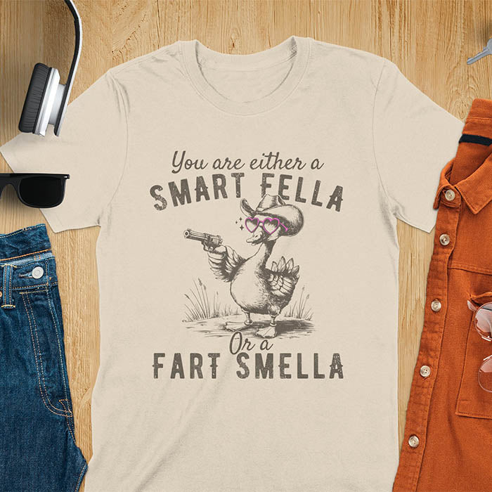 Sand color tee with A goose wearing heart shaped glasses and holding a pistol. Text reads You are either a Smart Fella or a Fart Smella.