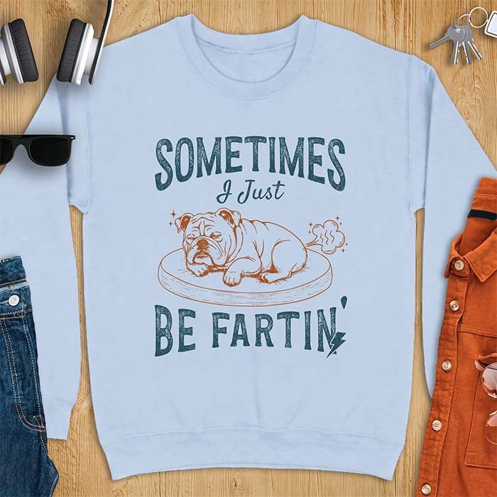 Light Blue sweatshirt with a graphic of a sleeping English Bulldog curled up on a dog bed with a fart cloud drawn behind it beside the text Sometimes I Just Be Fartin