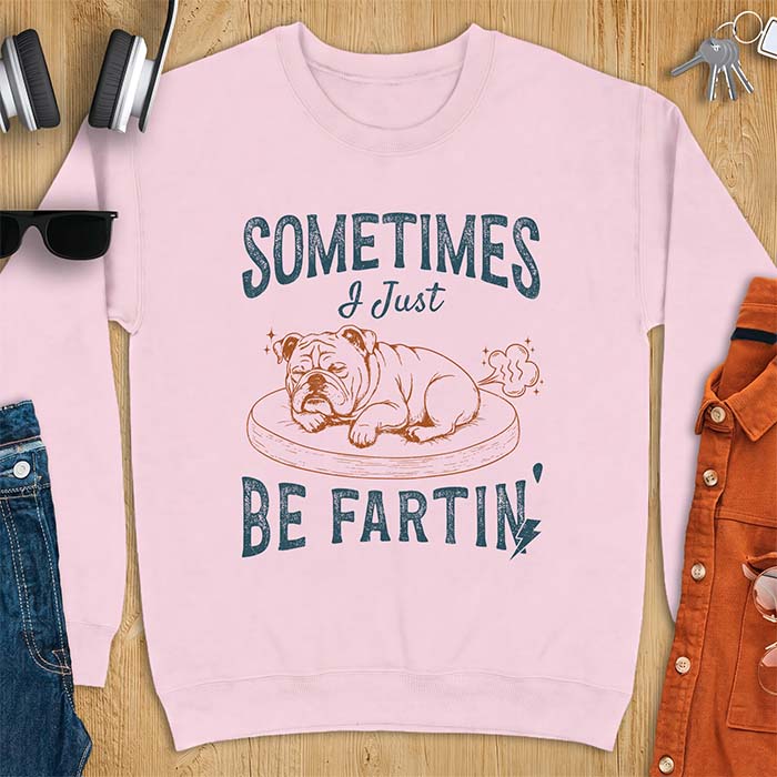 Light Pink sweatshirt with a graphic of a sleeping English Bulldog curled up on a dog bed with a fart cloud drawn behind it beside the text Sometimes I Just Be Fartin