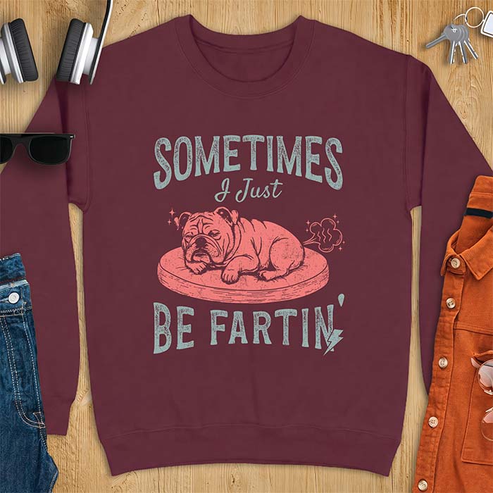 Maroon sweatshirt with a graphic of a sleeping English Bulldog curled up on a dog bed with a fart cloud drawn behind it beside the text Sometimes I Just Be Fartin