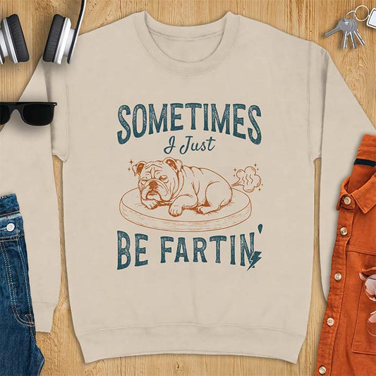 Sand sweatshirt with a graphic of a sleeping English Bulldog curled up on a dog bed with a fart cloud drawn behind it beside the text Sometimes I Just Be Fartin