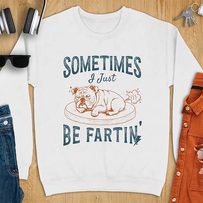 White sweatshirt with a graphic of a sleeping English Bulldog curled up on a dog bed with a fart cloud drawn behind it beside the text Sometimes I Just Be Fartin