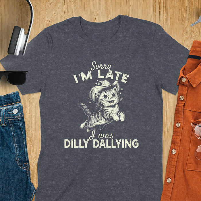 A dark heather t-shirt with a tabby cat wearing a cowboy hat. Text: Sorry I'M LATE above and I was DILLY DALLYING below. This cat lover shirt features cat cuteness gift and kawaiiness cuteness. A sarcastic meme shirt about dilly dallying, a funny genz meme with funny animals. This cowboy cat design has retro aesthetics, a snarky humorous tee, an animal person gift or mom gift.