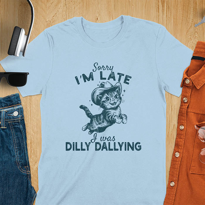 A light blue t-shirt with a tabby cat wearing a cowboy hat. Text: Sorry I'M LATE above and I was DILLY DALLYING below. This cat lover shirt features cat cuteness gift and kawaiiness cuteness. A sarcastic meme shirt about dilly dallying, a funny genz meme with funny animals. This cowboy cat design has retro aesthetics, a snarky humorous tee, an animal person gift or mom gift.