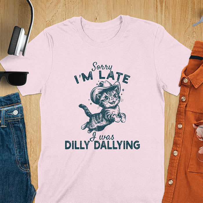 A light pink t-shirt with a tabby cat wearing a cowboy hat. Text: Sorry I'M LATE above and I was DILLY DALLYING below. This cat lover shirt features cat cuteness gift and kawaiiness cuteness. A sarcastic meme shirt about dilly dallying, a funny genz meme with funny animals. This cowboy cat design has retro aesthetics, a snarky humorous tee, an animal person gift or mom gift.