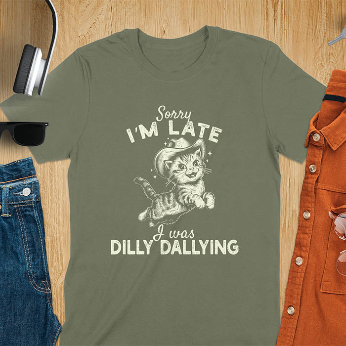 A military green t-shirt with a tabby cat wearing a cowboy hat. Text: Sorry I'M LATE above and I was DILLY DALLYING below. This cat lover shirt features cat cuteness gift and kawaiiness cuteness. A sarcastic meme shirt about dilly dallying, a funny genz meme with funny animals. This cowboy cat design has retro aesthetics, a snarky humorous tee, an animal person gift or mom gift.