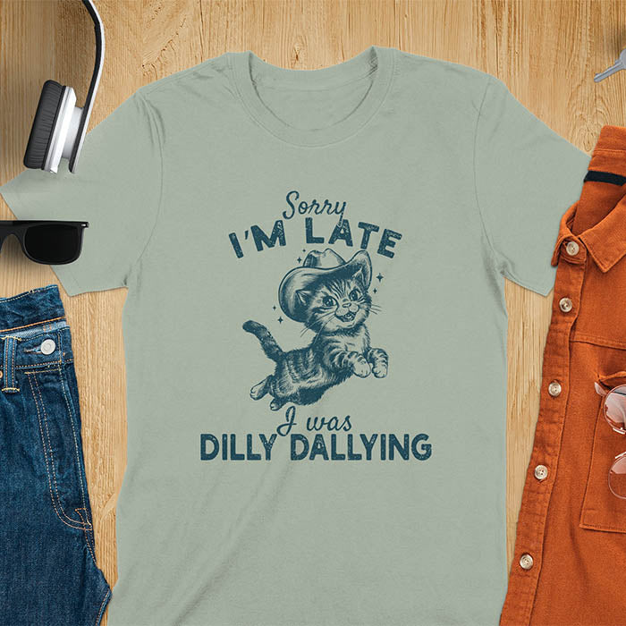 A sage t-shirt with a tabby cat wearing a cowboy hat. Text: Sorry I'M LATE above and I was DILLY DALLYING below. This cat lover shirt features cat cuteness gift and kawaiiness cuteness. A sarcastic meme shirt about dilly dallying, a funny genz meme with funny animals. This cowboy cat design has retro aesthetics, a snarky humorous tee, an animal person gift or mom gift.
