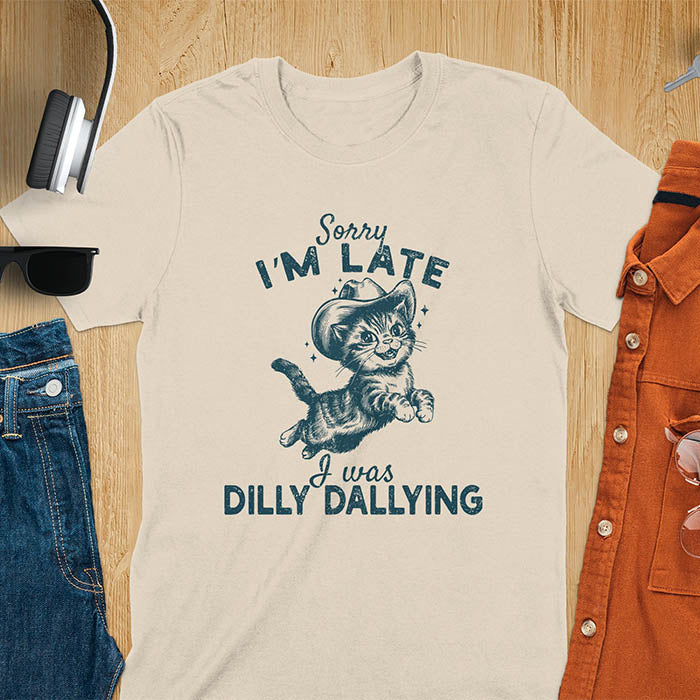 A sand color t-shirt with a tabby cat wearing a cowboy hat. Text: Sorry I'M LATE above and I was DILLY DALLYING below. This cat lover shirt features cat cuteness gift and kawaiiness cuteness. A sarcastic meme shirt about dilly dallying, a funny genz meme with funny animals. This cowboy cat design has retro aesthetics, a snarky humorous tee, an animal person gift or mom gift.