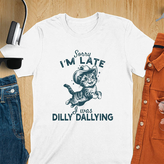 A white color t-shirt with a tabby cat wearing a cowboy hat. Text: Sorry I'M LATE above and I was DILLY DALLYING below. This cat lover shirt features cat cuteness gift and kawaiiness cuteness. A sarcastic meme shirt about dilly dallying, a funny genz meme with funny animals. This cowboy cat design has retro aesthetics, a snarky humorous tee, an animal person gift or mom gift.
