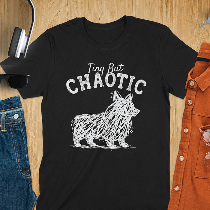 Black t-shirt featuring a corgi scribble line art graphic with the text "Tiny But Chaotic" on a wooden flat lay background