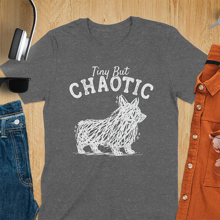 Dark heather t-shirt featuring a corgi scribble line art graphic with the text "Tiny But Chaotic" on a wooden flat lay background