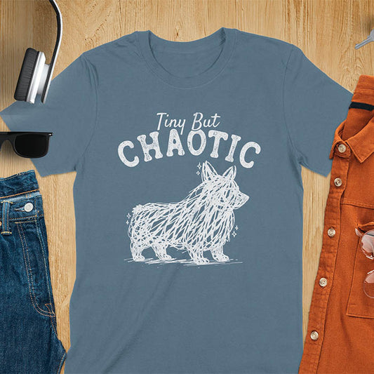 Indigo blue t-shirt featuring a corgi scribble line art graphic with the text "Tiny But Chaotic" on a wooden flat lay background