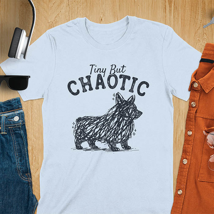 Light blue t-shirt featuring a corgi scribble line art graphic with the text "Tiny But Chaotic" on a wooden flat lay background