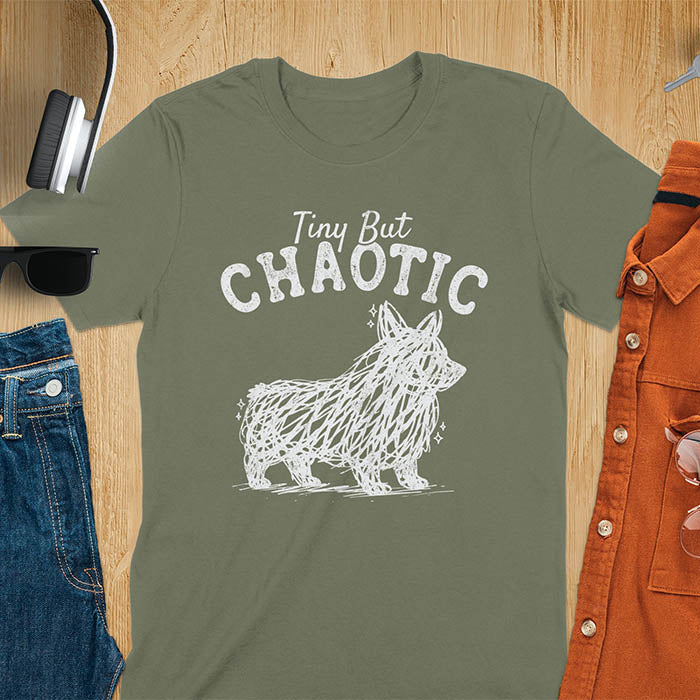 Military green t-shirt featuring a corgi scribble line art graphic with the text "Tiny But Chaotic" on a wooden flat lay background