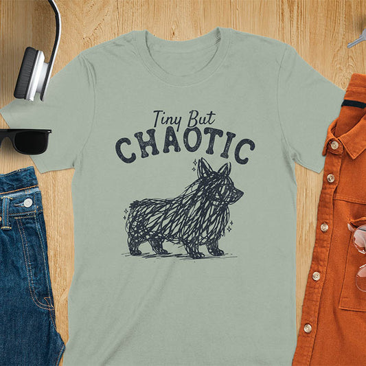 Sage green t-shirt featuring a corgi scribble line art graphic with the text "Tiny But Chaotic" on a wooden flat lay background