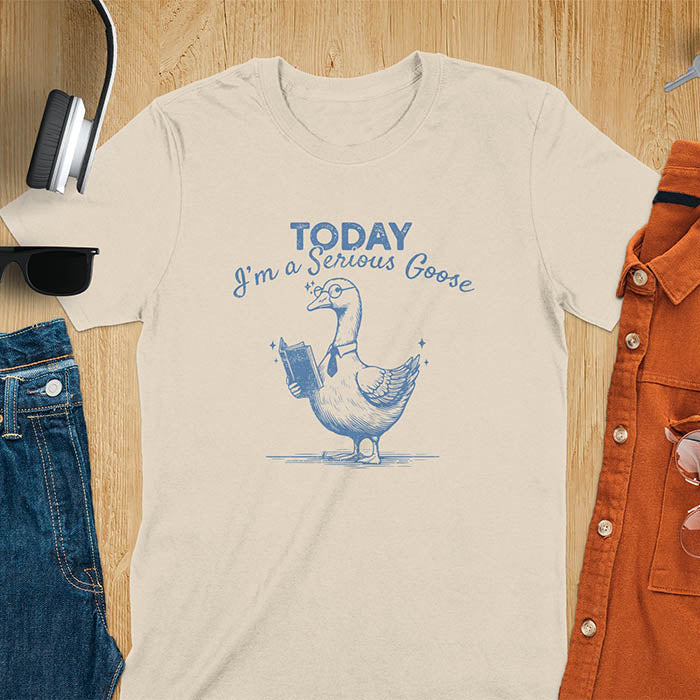 Sand color tee Featuring a retro vintage design with a distressed texture effect, this meme graphic tee showcases a dapper goose standing tall with a tie, collar, spectacles, and an open book in its wing, paired with the cheeky text, Today I’m a Serious Goose.