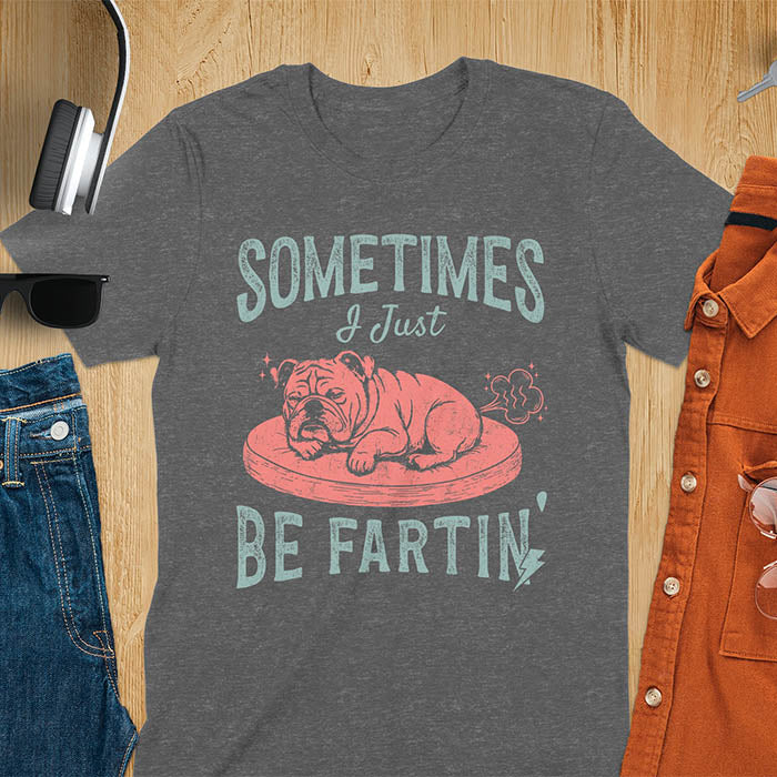 Dark heather t-shirt with vintage-inspired bulldog illustration. Retro graphic features snoozing English bulldog on dog bed with text 'Sometimes I Just Be Fartin' in distressed typography. Quirky animal meme design.