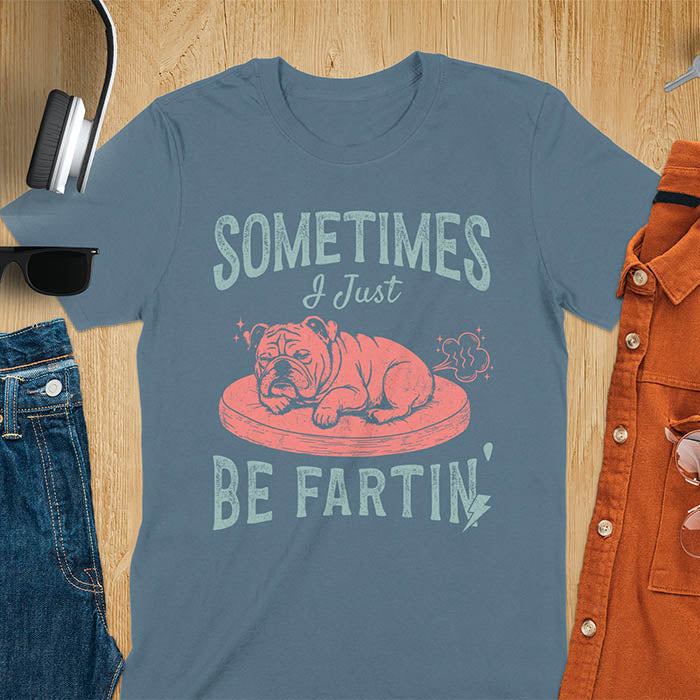 Indigo blue t-shirt with vintage-inspired bulldog illustration. Retro graphic features snoozing English bulldog on dog bed with text 'Sometimes I Just Be Fartin' in distressed typography. Quirky animal meme design.