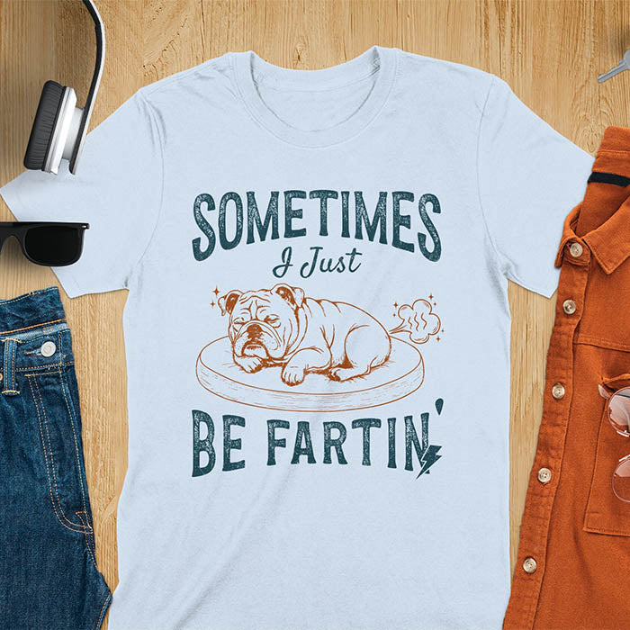 Light blue t-shirt with vintage-inspired bulldog illustration. Retro graphic features snoozing English bulldog on dog bed with text 'Sometimes I Just Be Fartin' in distressed typography. Quirky animal meme design.
