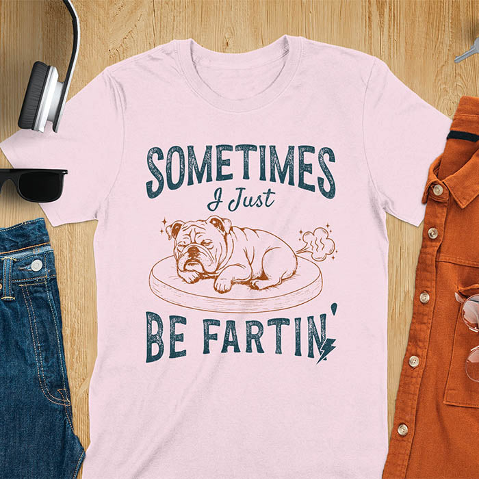 Light pink t-shirt with vintage-inspired bulldog illustration. Retro graphic features snoozing English bulldog on dog bed with text 'Sometimes I Just Be Fartin' in distressed typography. Quirky animal meme design.