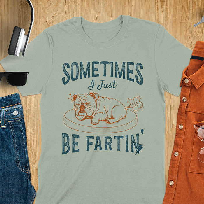 Sage green t-shirt with vintage-inspired bulldog illustration. Retro graphic features snoozing English bulldog on dog bed with text 'Sometimes I Just Be Fartin' in distressed typography. Quirky animal meme design.