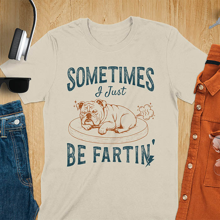 Sand color t-shirt with vintage-inspired bulldog illustration. Retro graphic features snoozing English bulldog on dog bed with text 'Sometimes I Just Be Fartin' in distressed typography. Quirky animal meme design. 