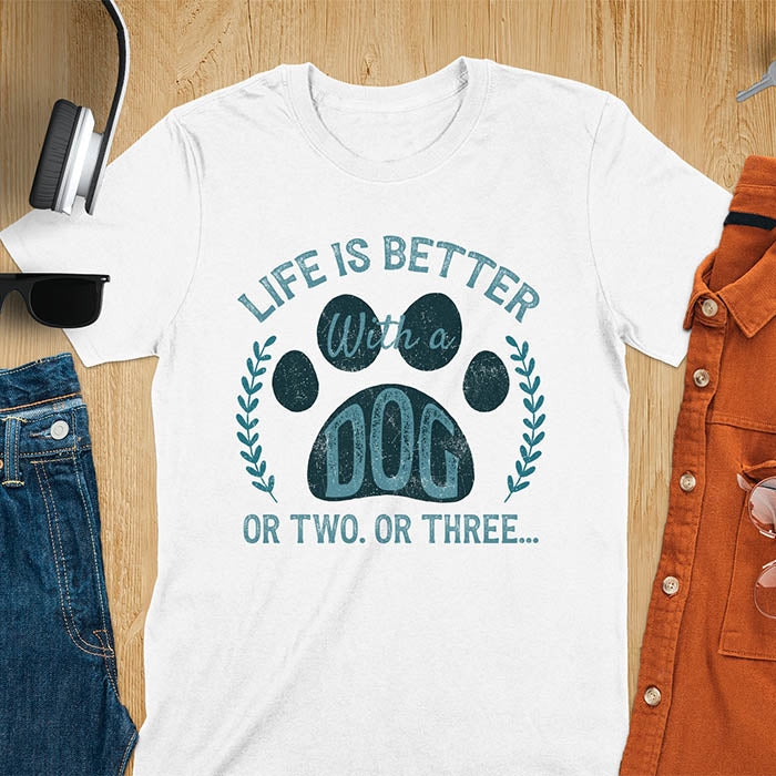 White t-shirt featuring large paw print with text inside surrounded by laurel wreath, text reads Life Is Better With A Dog Or Two Or Three